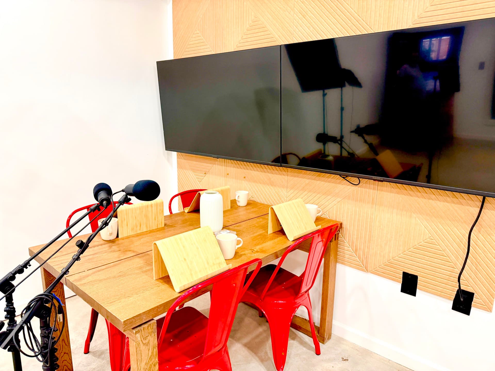 The podcast studio: a wooden recording table with red chairs, microphones on boom arms and mugs set out, beneath two wall-mounted screens.